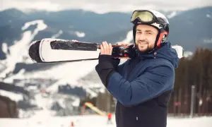 Uśmiechnięty narciarz w kasku i goglach niesie na ramieniu narty dla średniozaawansowanych. W tle ośnieżone góry.