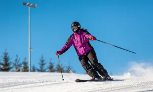 Narciarka w fioletowej kurtce zjeżdża po stoku, wzbijając śnieg.
