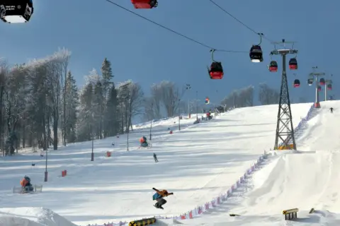 Kolejka gondolowa nad ośnieżonym stokiem narciarskim, na którym snowboardzista wykonuje skok.