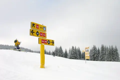 Żółte tablice z kolorami tras narciarskich wskazują drogę. W tle śnieżny stok, drzewa i armatka śnieżna.