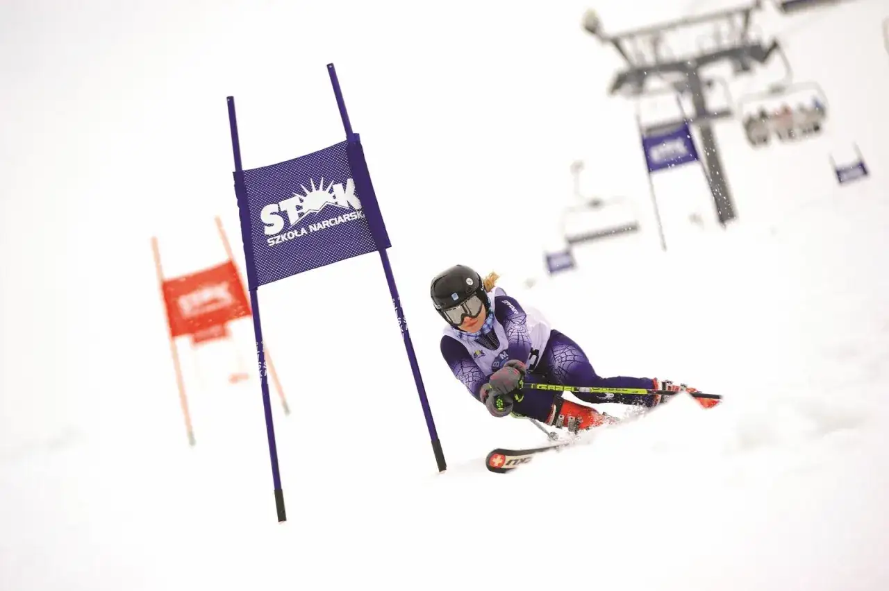 Narciarka wykonuje dynamiczny narty cross turn, mijając fioletową tyczkę z logo szkoły narciarskiej.