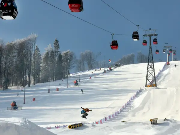 Kolejka gondolowa nad ośnieżonym stokiem narciarskim, na którym snowboardzista wykonuje skok.
