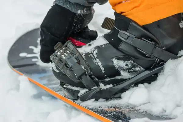 Rękawica zapina but snowboardowy na desce, przygotowując się do wygrzewania butów snowboardowych w śniegu.
